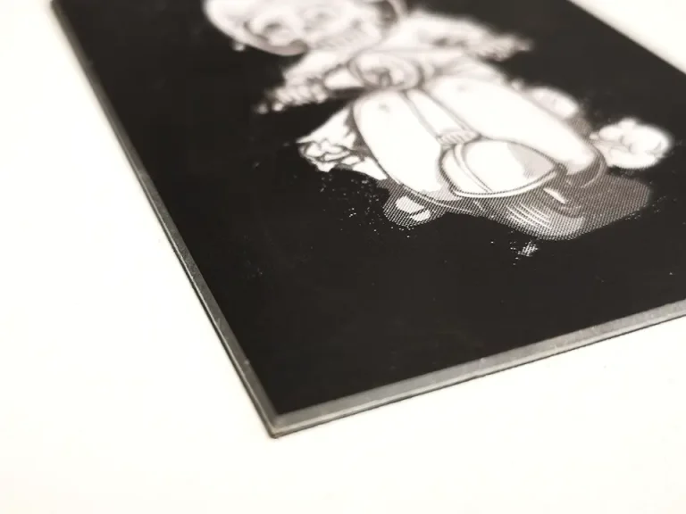 Gros plan sur le coin d'une carte rectangulaire noire, travaillée à la biseauteuse pour des bords lisses, comportant une illustration blanche d'un personnage stylisé aux grands yeux, assis. La carte repose sur une surface blanche.
