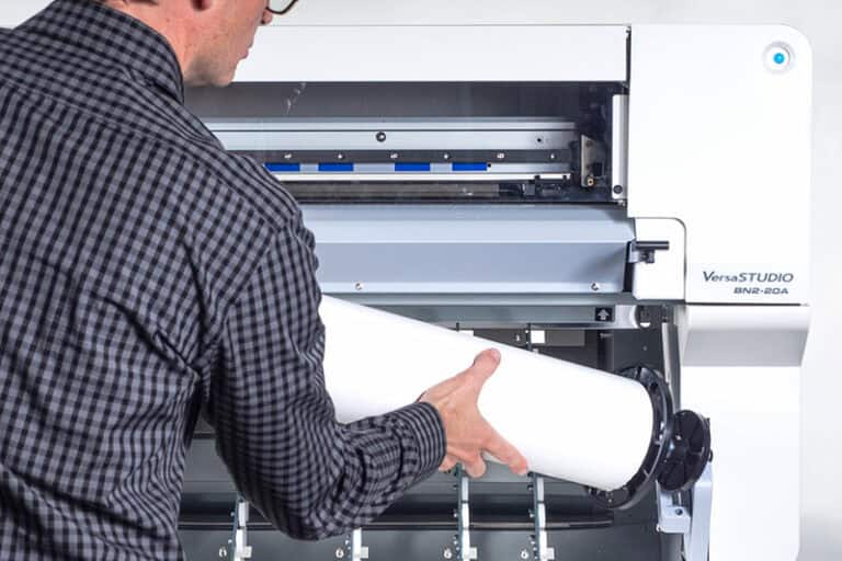 Une personne en chemise à carreaux insère un rouleau de papier dans une VersaSTUDIO BN-20A blanche, une imprimante/découpeuse à jet dencre. Le panneau avant de la machine est ouvert, révélant son mécanisme interne.