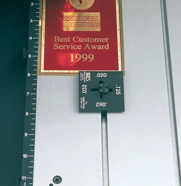 Une plaque avec le « American Express Best Customer Service Award 1999 » à côté dune jauge de mesure avec différents réglages dépaisseur, montée sur un mur avec une règle verticale à côté.