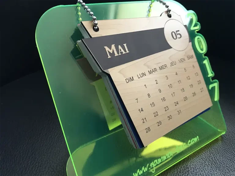 Calendrier de bureau sur support acrylique vert fluo indiquant le mois de mai 2017 en français. La date actuelle mise en évidence est le 5. Il est élégamment suspendu par des chaînes à billes en métal.