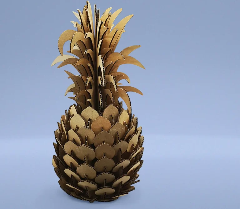 Sculpture en carton dun ananas sur fond bleu uni, réalisée avec découpe du carton au laser. La sculpture présente des couches et des détails complexes, imitant parfaitement la texture et la forme dun véritable ananas.