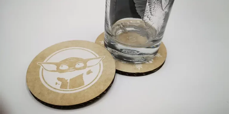 Un verre deau est posé sur un dessous de verre rond avec une illustration doiseau réalisée par découpe au laser. À côté, un autre dessous de verre présente un personnage de dessin animé avec de grandes oreilles et de grands yeux sur un fond uni. Tous deux sont fabriqués dans un matériau marron clair, soulignant la précision de la découpe au laser.