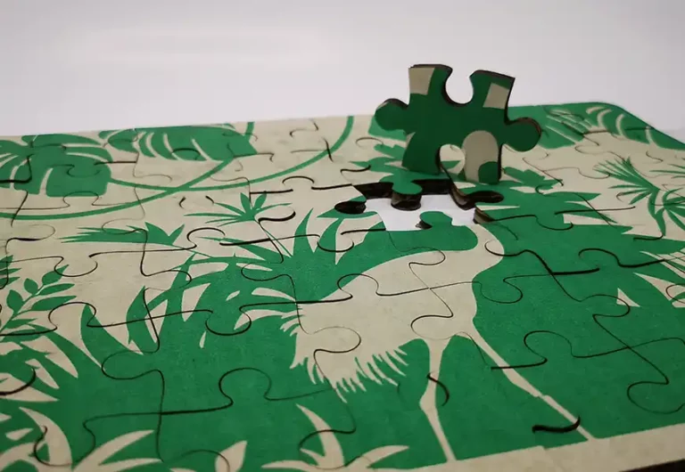Un puzzle en carton partiellement terminé, gravé et découpé au laser, présente un motif de plante tropicale verte et beige. Une pièce est soulevée, prête à sinsérer dans son emplacement vide sur le plateau du puzzle.