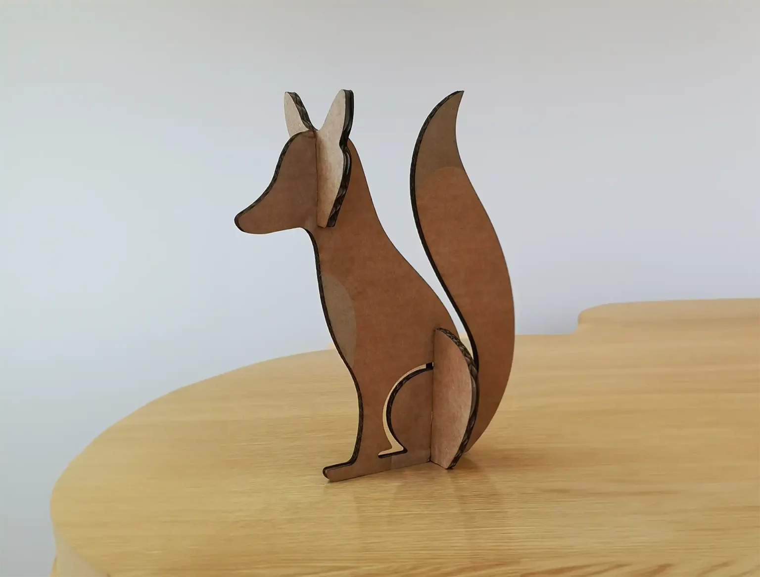 Une simple découpe en carton d'un renard aux oreilles pointues et à la queue touffue, découpée au laser, repose sur une surface en bois sur un fond neutre.