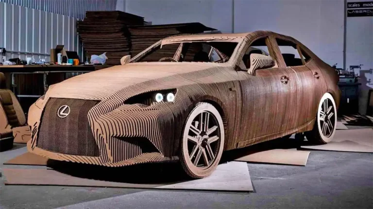 Une voiture grandeur nature entièrement fabriquée en carton ondulé, présentant des détails complexes et une précision de découpe au laser. Elle se trouve dans un atelier entouré doutils et de feuilles de carton, avec des phares et des roues réalistes mettant en valeur son design.