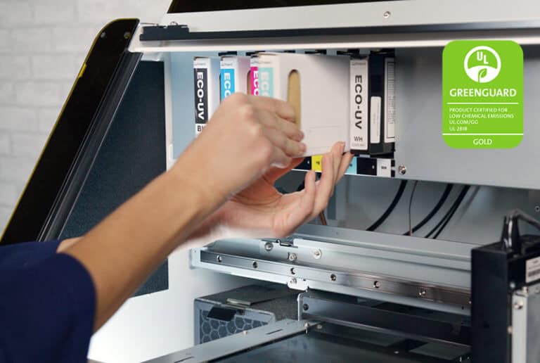 Une personne installe une cartouche dencre blanche dans une imprimante, créant ainsi une configuration impressionnante pour une impression de haute qualité. Lintérieur de la machine, conçu pour plusieurs cartouches, est clairement visible. Un emblème de certification UL GREENGUARD garantit un fonctionnement respectueux de lenvironnement dans le coin supérieur droit.