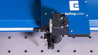 Gros plan dune machine laser Epilog bleue équipée dun système à double source en fonctionnement. Le bras mécanique plane précisément au-dessus de la surface de la grille, prêt à graver ou à découper avec une précision remarquable.