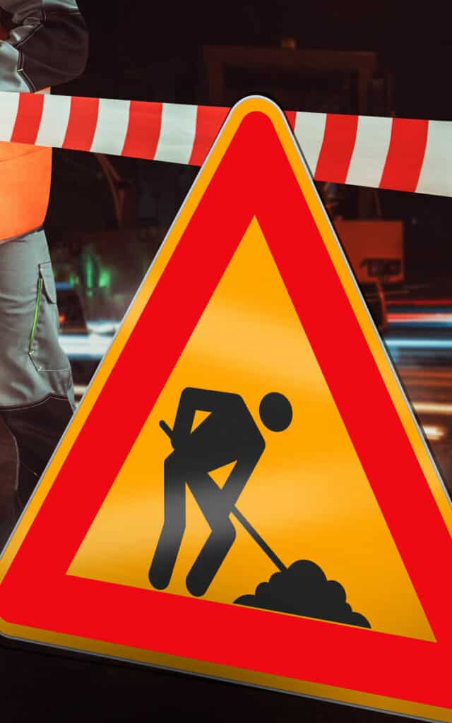 Un panneau d'avertissement triangulaire jaune et rouge pour la construction, probablement fabriqué avec un traceur de découpe, montre une silhouette noire creusant devant une barrière rayée rouge et blanche. Une partie d'un ouvrier vêtu d'un gilet orange est visible à gauche.