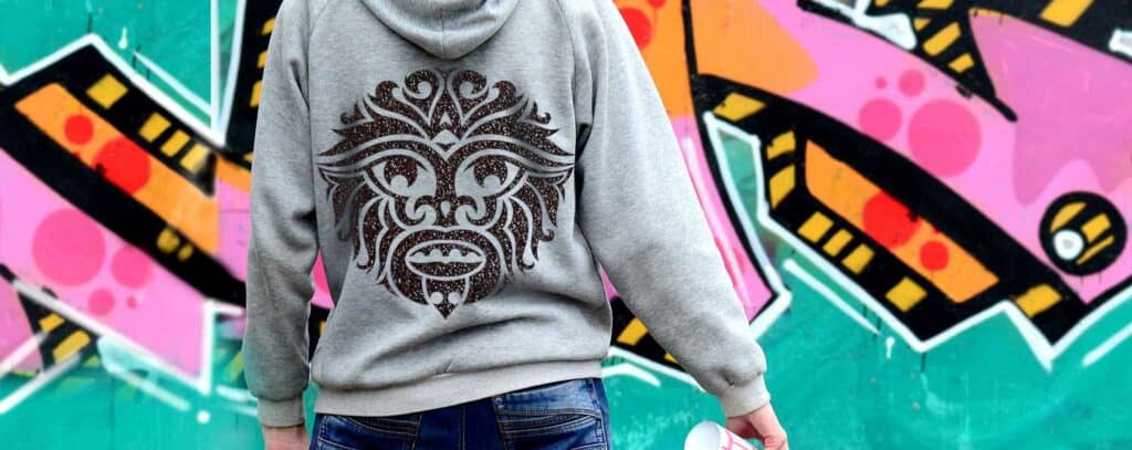 Une personne portant un sweat à capuche gris avec un motif de masque tribal dans le dos se tient près dun mur de graffitis vibrant, une bombe aérosol et un traceur de découpe à la main, créant de lart abstrait avec des tons roses, orange et verts.