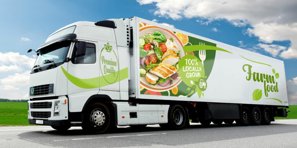 Un semi-remorque blanc au graphisme vert, créé à l'aide d'un traceur de découpe, porte les mentions "Farm food" et "100% local grown", avec une grande image d'une salade au poulet sur une route de campagne sous un ciel bleu.