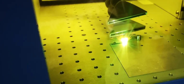 Vue rapprochée d'une machine de gravure laser à fibre galvaudée en fonctionnement, émettant une lumière verte-jaune brillante sur un matériau rectangulaire plat placé sur une surface métallique perforée.