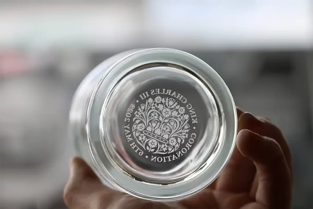 Une main présentant un verre héliogravure UV, tenu à lenvers pour révéler une inscription indiquant « ROI CHARLES III · COURONNEMENT · 2023 » avec un emblème décoratif. Larrière-plan flou ajoute un air de mystère.
