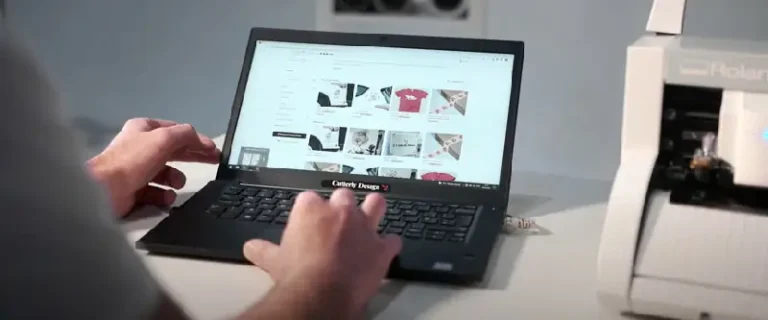 Une personne utilise un ordinateur portable pour afficher une boutique en ligne proposant divers vêtements et accessoires ; un traceur de découpe blanc est partiellement visible sur le côté droit du bureau.