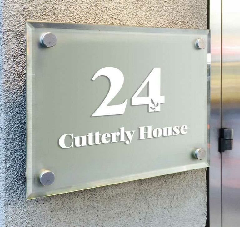Une enseigne en verre montée sur un mur gris affiche le numéro « 24 » et le nom « Cutterly House » en texte blanc, réalisés de main de maître avec un plotter de découpe. Lenseigne est fixée par des entretoises métalliques élégantes à chaque coin.