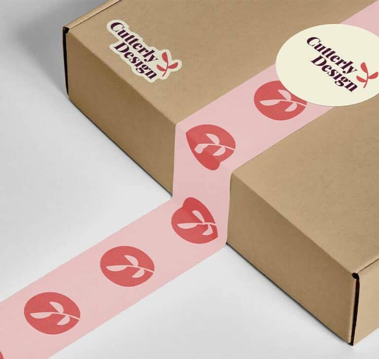 Une boîte en carton fermée par du ruban adhésif rose avec des illustrations de cerises rouges est présentée, ce qui laisse deviner la précision de sa fabrication. La boîte porte un autocollant « Cuttery Design », mettant en valeur le nom de la marque, probablement créé à laide dun plotter de découpe pour des détails complexes.