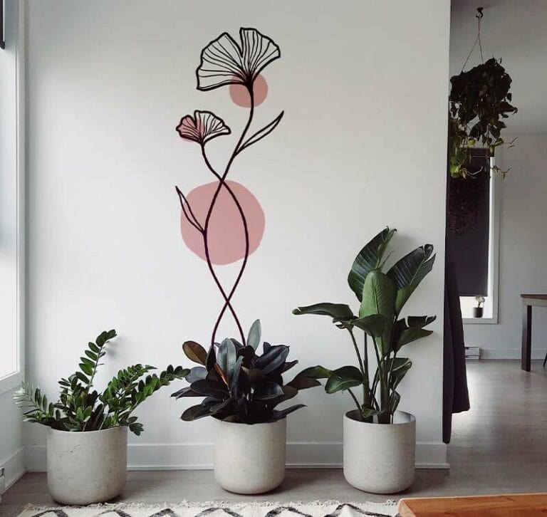 Chambre minimaliste avec de grandes œuvres dart florales abstraites réalisées à laide dun plotter de découpe, représentant deux fleurs et des cercles roses. Trois plantes en pot aux formes de feuilles variées sont posées sur un plancher en bois clair. Une grande fenêtre sur la gauche fournit de la lumière naturelle.