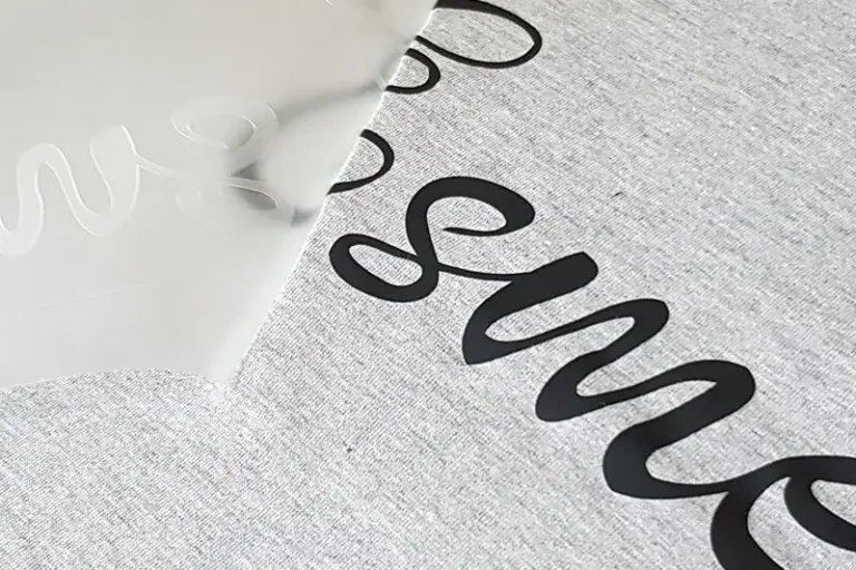 Un gros plan sur une surface grise révèle un lettrage en vinyle noir, minutieusement réalisé à laide dun plotter de découpe, partiellement recouvert dune feuille de transfert transparente. Le mot commence par « sm » dans une élégante police cursive, le reste étant masqué par la feuille.