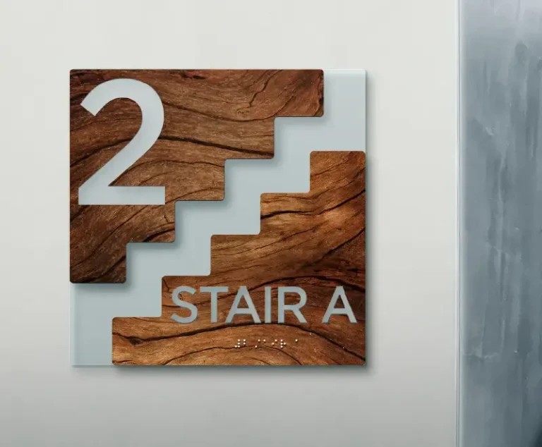 Un panneau sur un mur indique "2 STAIR A" en grosses lettres avec du braille en dessous. Réalisée en bois et en verre, l'enseigne - avec son graphisme d'escalier - a été imprimée sur objet pour indiquer l'escalier du deuxième étage.