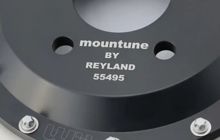 Gros plan dune pièce métallique noire avec la gravure complexe « mountune BY REYLAND 55495 », démontrant une habileté experte dans la gravure du métal. La conception circulaire comporte des trous de boulon disposés sur le pourtour, mettant en valeur à la fois la forme et la fonction.