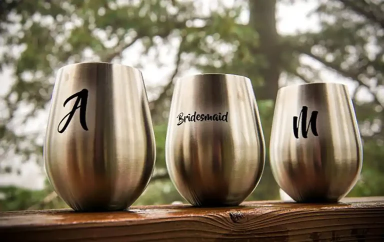 Trois verres à vin en acier inoxydable sur un support en bois, chacun gravé avec soin dun texte différent. Le premier porte un « A », celui du milieu porte linscription « Bridesmaid » et le troisième un « M ». Les arbres sont flous en arrière-plan, mettant en valeur lart de graver le métal.