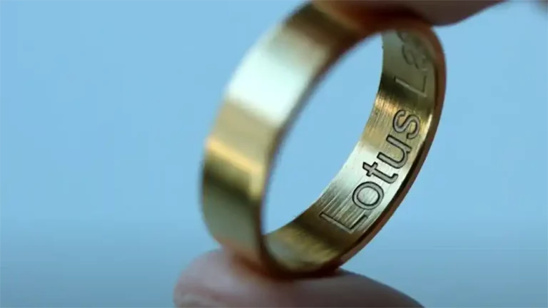 Gros plan dune bague en or, gravée avec brio, tenue entre deux doigts. Le mot Lotus est élégamment gravé à lintérieur. Larrière-plan se brouille doucement dans des tons bleus.