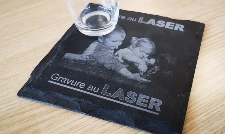 Un verre repose sur un élégant dessous de verre en ardoise noire orné dune image gravée au laser de deux enfants. Linscription se lit Gravure au LASER en blanc. Cette belle pièce met en valeur lélégance de la gravure au laser sur ardoise, complétant à merveille son support en bois.
