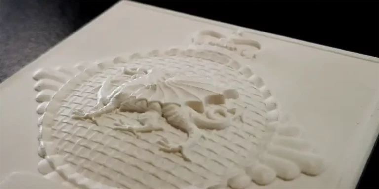 Gros plan dun dragon texturé imprimé en 3D au motif ovale, rappelant la gravure au laser sur ardoise, entouré dun motif tissé complexe et couronné au sommet, le tout en blanc cassé monochrome. Léclairage met en valeur les détails en relief.