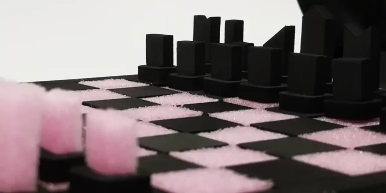 Gros plan d'un échiquier avec des cases roses et des pièces noires découpées au laser de forme unique, placées de manière stratégique. Le fond blanc uni renforce le contraste saisissant entre les éléments roses et noirs.