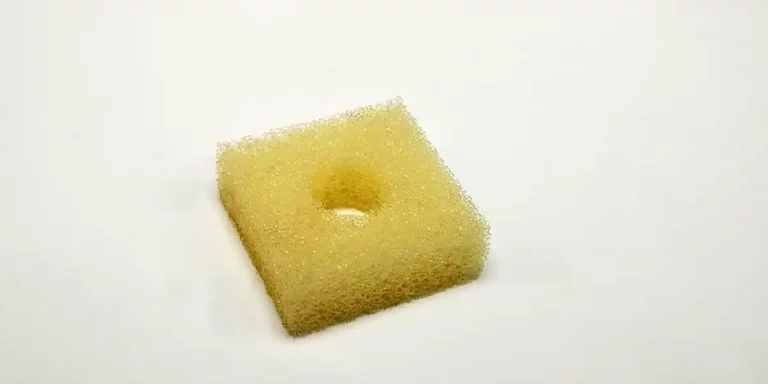 Une éponge carrée jaune avec un trou découpé au laser au centre repose sur un fond blanc uni.