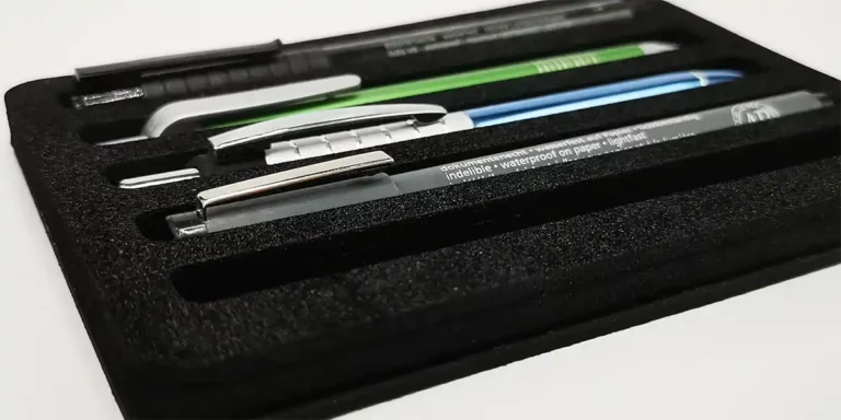 Un étui en mousse noire, découpé avec précision au laser, contient une variété de stylos et de crayons. Lensemble comprend des stylos aux corps bleu, vert et noir, chacun niché dans sa propre fente pour un rangement organisé. Limage présente létui ouvert sous un angle.
