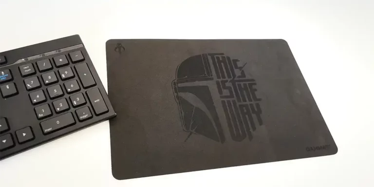 Un clavier noir est partiellement visible à côté dun tapis de souris noir avec un motif de casque et le texte « This Is The Way », réalisé avec précision à laide de techniques de découpe laser mousse.