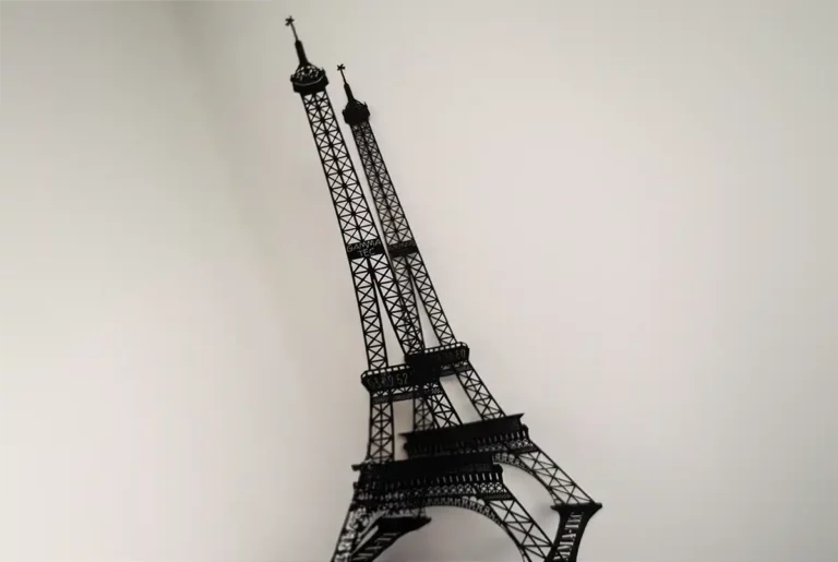 Une maquette décorative en métal noir de la Tour Eiffel se détache sur un fond blanc uni, projetant une ombre vers la gauche. Sa structure en treillis complexe, rappelant la découpe et la gravure du papier, ressemble magnifiquement à lemblématique monument parisien.
