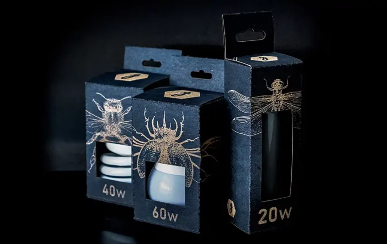 Trois boîtes en carton bleu foncé avec des illustrations dinsectes découpées et gravées sur papier, chacune abritant une ampoule étiquetée avec différentes puissances : 40 W, 60 W et 20 W. Les boîtes présentent un design moderne et sont astucieusement disposées sur un fond noir.