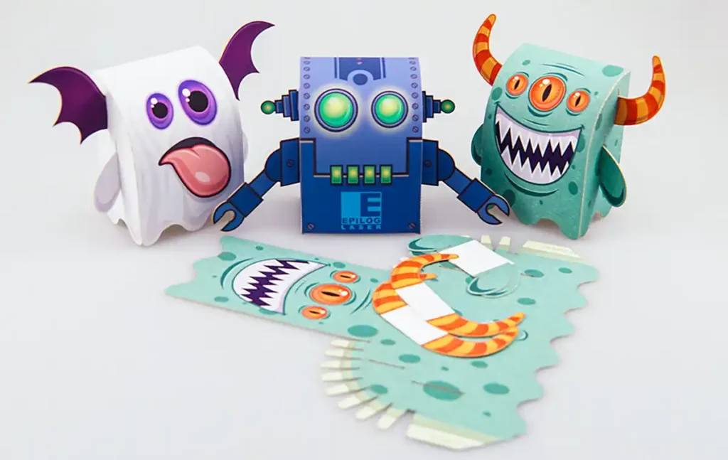 Trois personnages en papier coloré, réalisés selon lart de la découpe et de la gravure du papier, sont disposés sur une surface. Le robot bleu a des yeux brillants, le fantôme est blanc avec des ailes violettes et le monstre vert possède des cornes et une bouche ouverte.