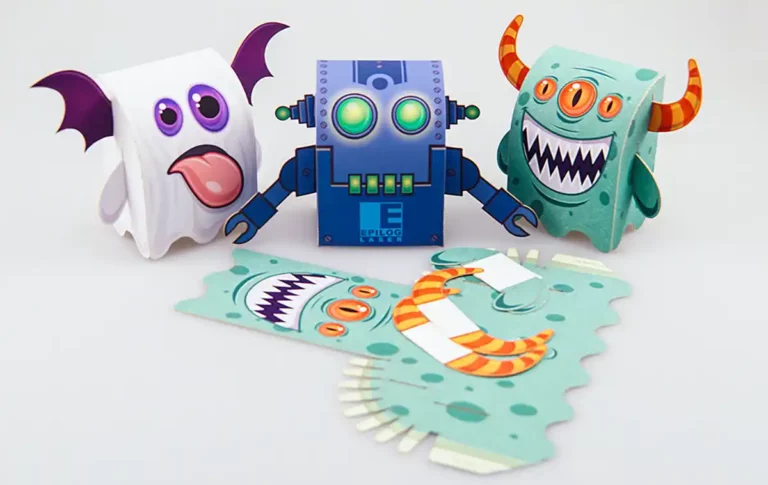 Trois personnages en papier coloré, réalisés selon lart de la découpe et de la gravure du papier, sont disposés sur une surface. Le robot bleu a des yeux brillants, le fantôme est blanc avec des ailes violettes et le monstre vert possède des cornes et une bouche ouverte.