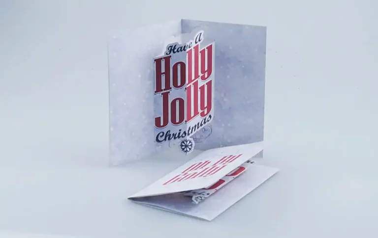 Une carte de vœux pliée sur fond blanc affiche le message « Have a Holly Jolly Christmas » en lettres rouges et noires. Dotée dune découpe et dune gravure sur papier, la carte est légèrement ouverte, révélant une carte finement conçue en dessous.