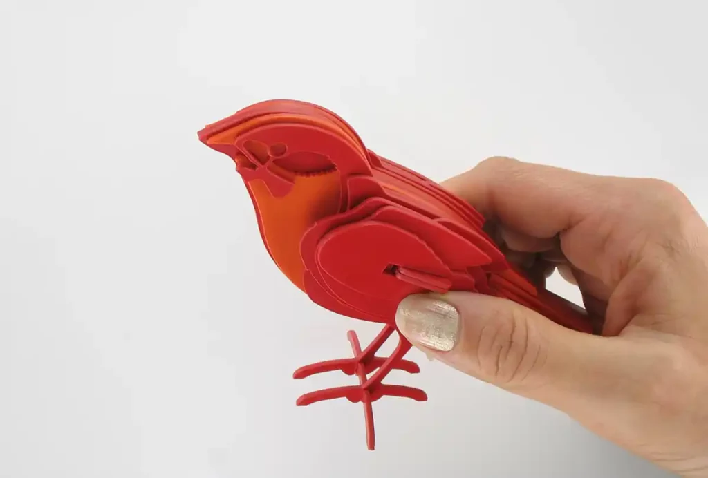 Une personne aux ongles peints tient un modèle doiseau rouge imprimé en 3D avec des sections superposées, mettant en valeur la découpe et la gravure expertes du plastique, sur un fond blanc.