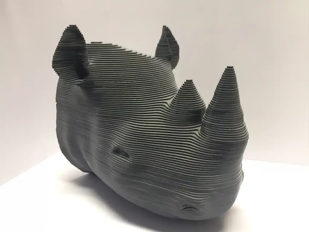 Sculpture en couches dune tête de rhinocéros réalisée à partir de plaques horizontales noires empilées et régulièrement espacées sur un fond uni. Le motif met en valeur les cornes et les traits du visage du rhinocéros avec une apparence moderne et abstraite, rappelant les techniques de découpe et de gravure du plastique.