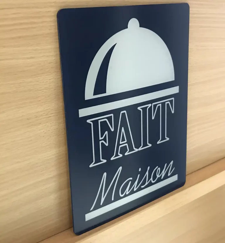 Une enseigne noire et blanche présentant un motif de cloche avec les mots « Fait Maison » bien en évidence, réalisée de main de maître par découpe et gravure du plastique, placée sur un fond de bois clair.