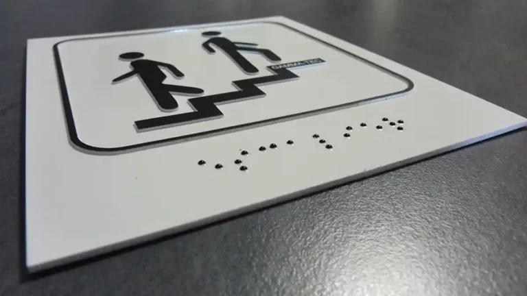 Un panneau tactile, réalisé de main de maître par découpe et gravure du plastique, présente des symboles en relief de deux personnages sur des escaliers ainsi quun texte en braille. Le panneau est monté sur une surface grise.