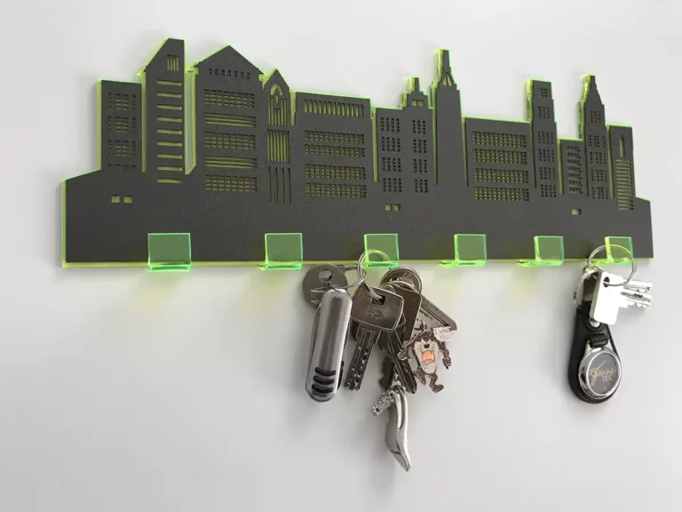 Un porte-clés en acrylique conçu comme une silhouette de skyline de ville avec des accents verts. Trois jeux de clés sont suspendus au support, chacun sur un crochet séparé. La skyline présente différentes formes de bâtiments avec des fenêtres découpées.
