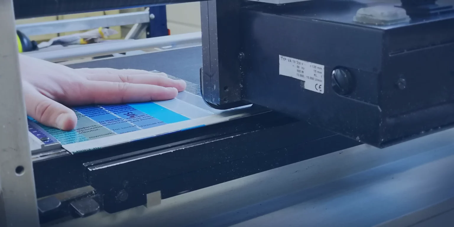 La main dune personne guide avec brio une feuille de papier, présentant les finitions des matières, dans une machine à imprimer. Le papier porte un motif déchantillons de couleurs vives alors que la machine fonctionne dans un environnement industriel.