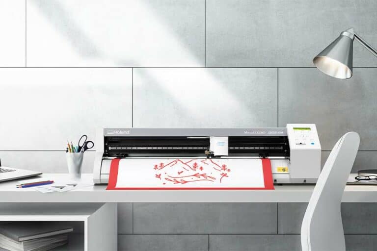 Un espace de travail moderne est doté dun plotter de découpe sur le bureau, découpant avec précision un graphique rouge de montagnes et darbres. Linstallation comprend une lampe, un porte-papeterie et un ordinateur portable, le tout posé contre un élégant mur carrelé gris.