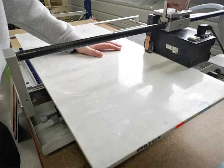 Une personne utilise une machine à découper les carreaux, en appuyant sur un grand carreau blanc placé sur la plate-forme du coupeur. La main maintient le carreau tandis que la lame - semblable à une scie circulaire varga - est positionnée de manière à effectuer une coupe droite.