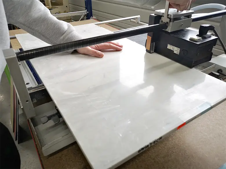 Une personne utilise une machine à découper les carreaux, en appuyant sur un grand carreau blanc placé sur la plate-forme du coupeur. La main maintient le carreau tandis que la lame - semblable à une scie circulaire varga - est positionnée de manière à effectuer une coupe droite.