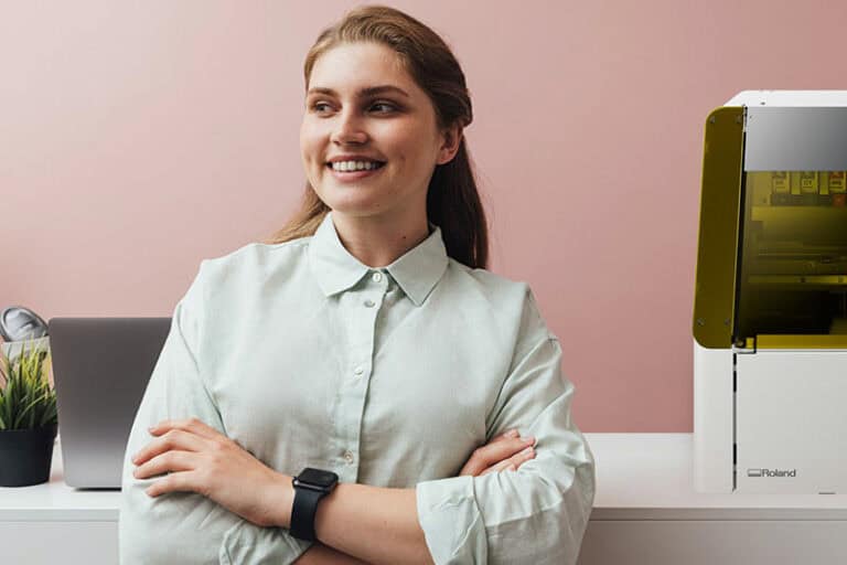 Une personne aux cheveux longs sourit, les bras croisés, en chemise claire et montre. Ils sont à côté dun ordinateur portable et dune machine à impression objet sur une table blanche avec une plante en pot à proximité. Le mur rose pâle offre la toile de fond parfaite pour cette scène sereine.