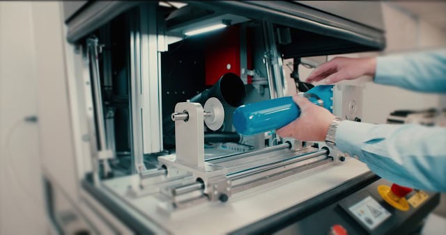Une personne place soigneusement un objet cylindrique bleu dans un cadre métallique à lintérieur de la machine. Cette pièce complexe, qui fait probablement partie dun processus de gravure industrielle, est dotée de divers composants mécaniques et dun panneau de commande qui brille sous la précision de la technologie laser.