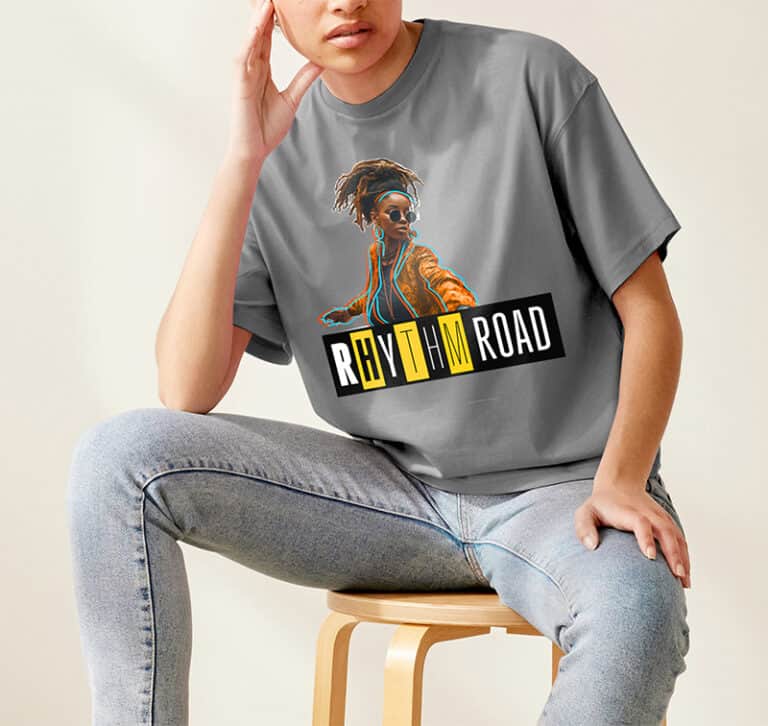 Une personne assise sur un tabouret porte un t-shirt gris avec un imprimé à jet dencre représentant une femme élégante et le texte « Rhythm Road », associé à un jean bleu clair. La personne pose son coude sur son genou, dans une pose détendue sur un fond uni.