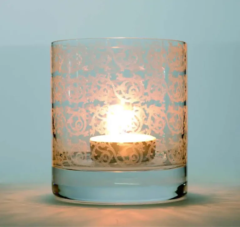 Une bougie chauffe-plat allumée dans un support en verre transparent avec un motif floral complexe et givré réalisé à laide de verre héliogravure UV. La lueur chaleureuse de la bougie illumine le design, créant une ambiance douce et chaleureuse sur un fond clair et neutre.