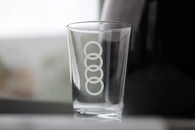 Un verre transparent orné de quatre cercles blancs entrelacés, créés par gravure verre UV, se dresse élégamment sur une surface réfléchissante sur un fond gris et blanc flou.